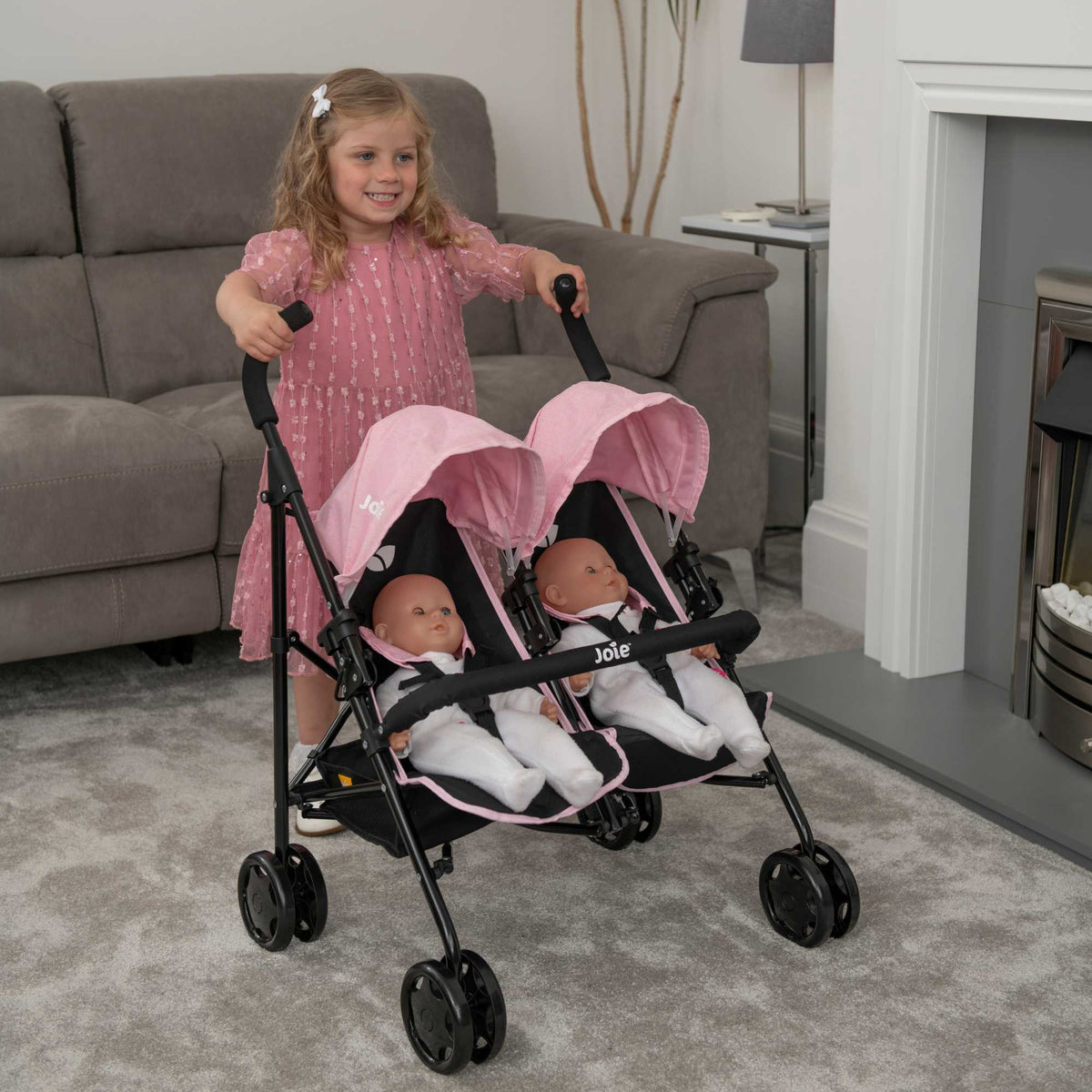 Joie Aire Twin Dolls Stroller in pink and black, designed with side-by-side seating for two dolls, adjustable handle height, swivel wheels, and a large storage basket, ideal for children to enjoy twin doll outings.