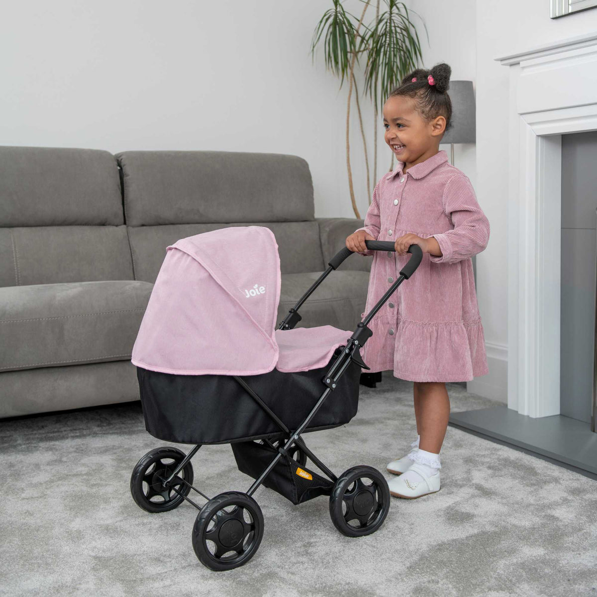 Joie Baby Dolls Pram in pink and black, featuring adjustable handle height, swivel wheels, and a spacious storage basket, designed for children to enjoy imaginative play with their dolls.