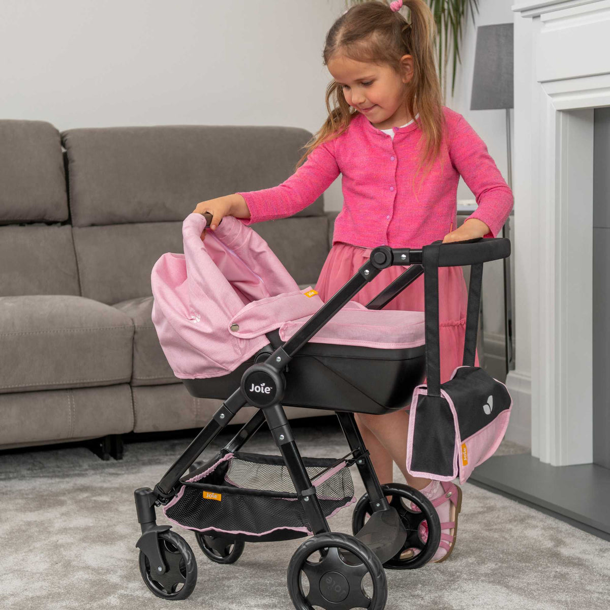 Joie Mytrax Dolls Pram in pink and black, featuring a realistic design with adjustable handle height, swivel wheels, and a large storage basket, ideal for children to take their dolls on walks and play pretend.