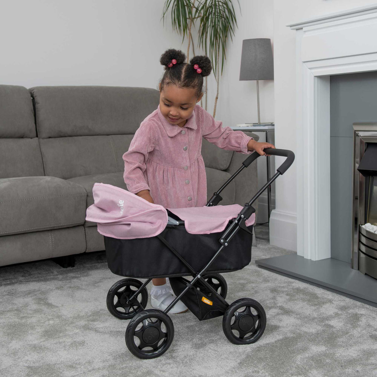 Joie Baby Dolls Pram in pink and black, featuring adjustable handle height, swivel wheels, and a spacious storage basket, designed for children to enjoy imaginative play with their dolls.