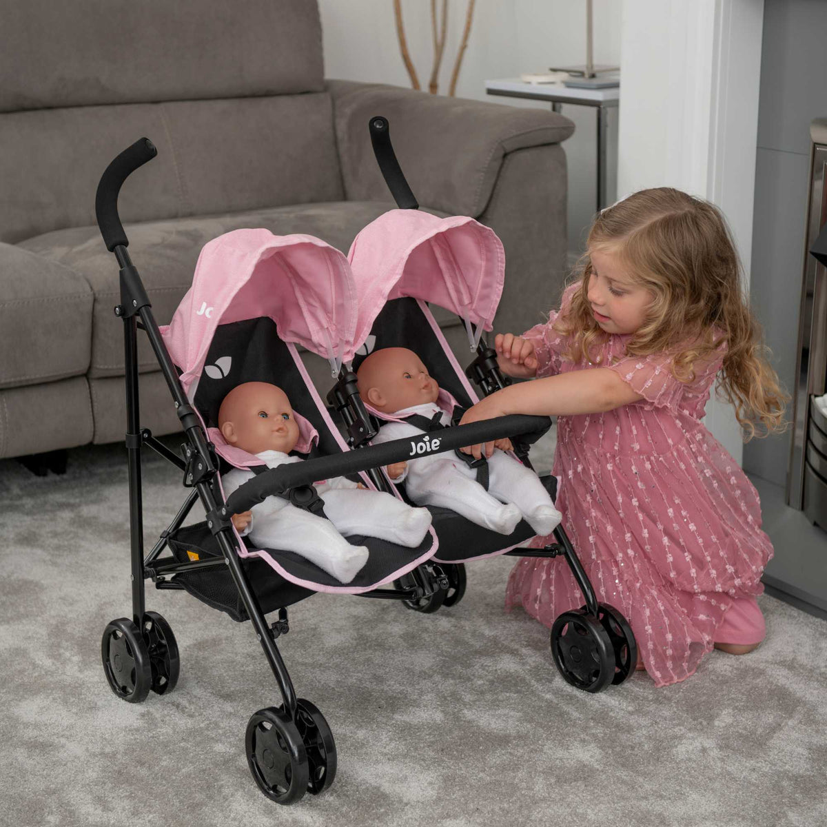 Joie Aire Twin Dolls Stroller in pink and black, designed with side-by-side seating for two dolls, adjustable handle height, swivel wheels, and a large storage basket, ideal for children to enjoy twin doll outings.